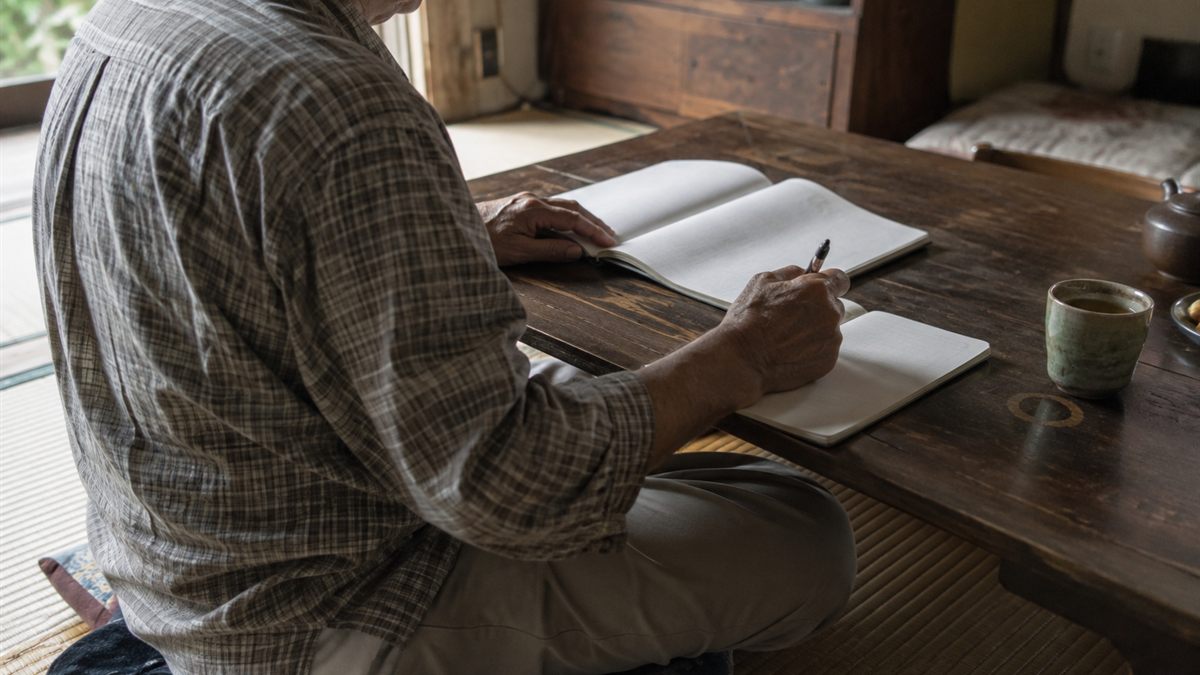 低い座卓で冊子を開き、手元のノートに書き留めている場面